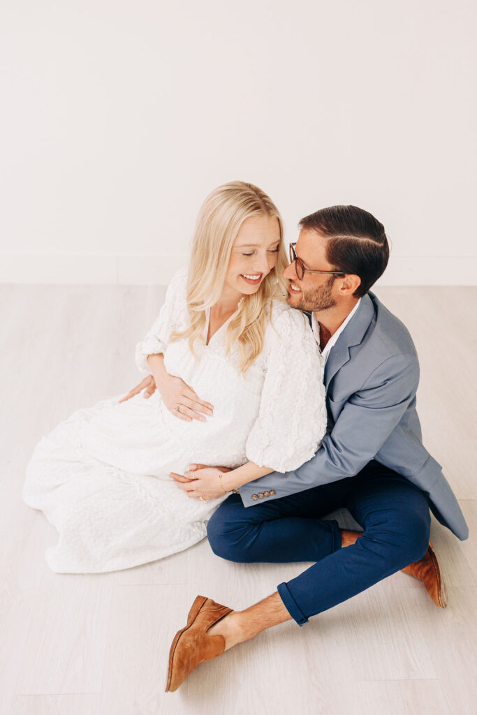 A mom to be leans into her husband while sitting on the floor of a studio in a white maternity gown prenatal massage in houston