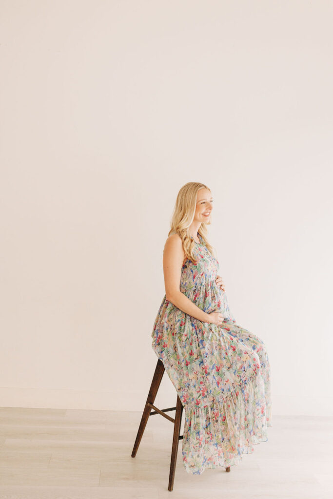 A mom to be in a colorful maternity dress sits on a wooden stool prenatal massage in houston
