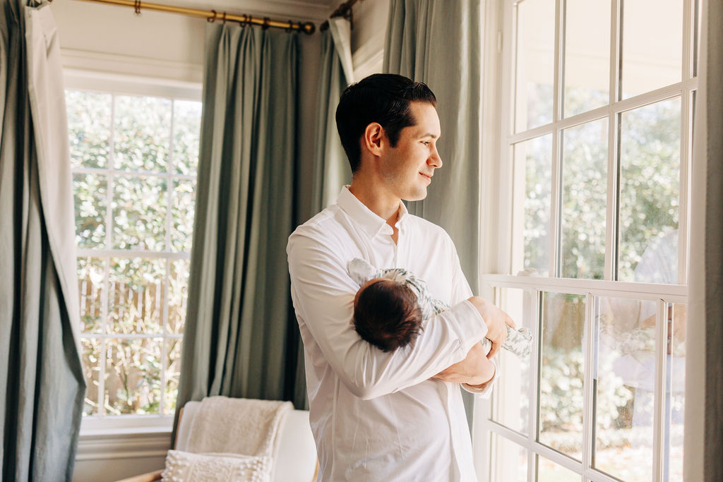 A new father gazes out a window while holding his sleeping newborn baby spas in houston