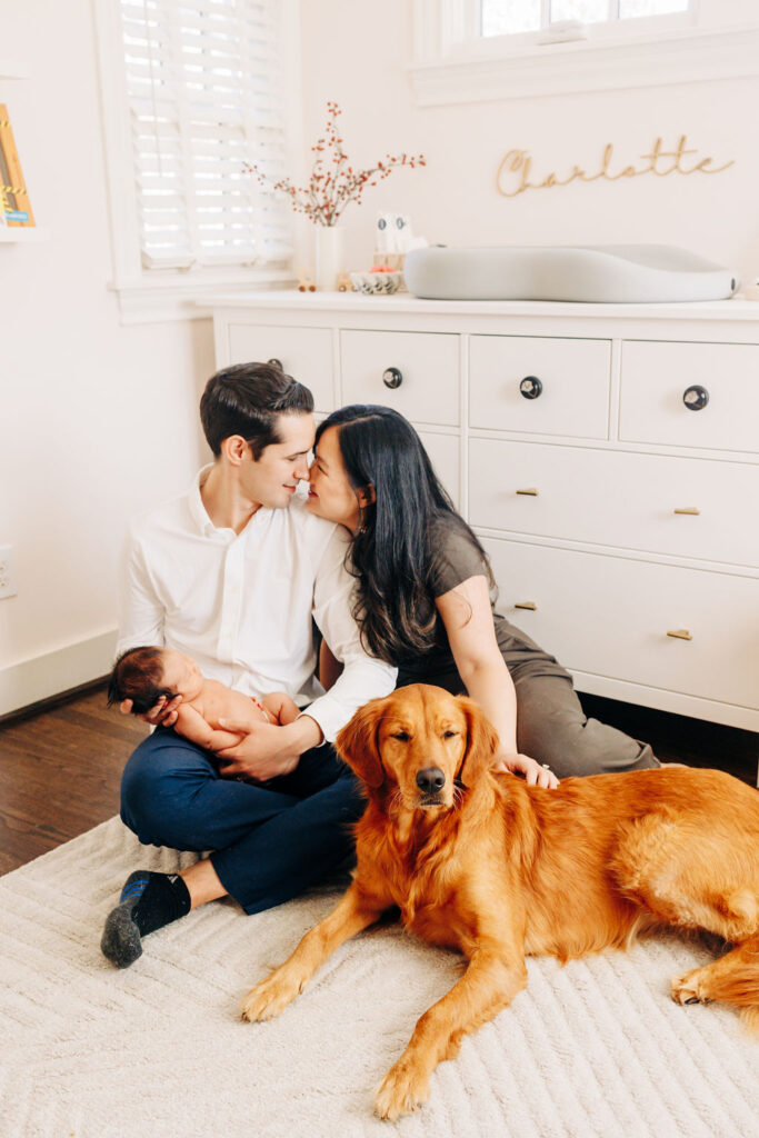 New parents kiss while sitting on their nursery floor holding their sleeping newborn baby with their golden retriever spas in houston