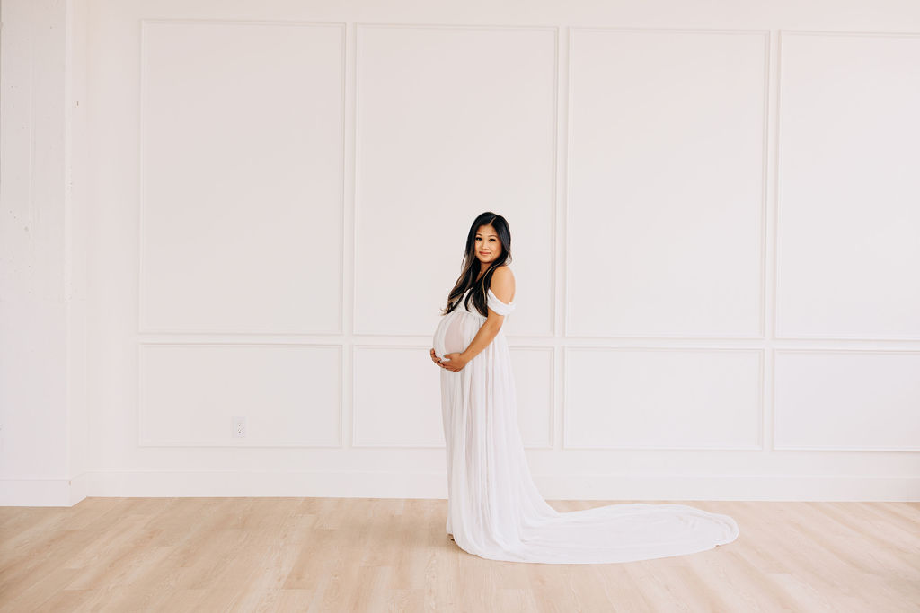A mom to be in a white maternity gown stands in a studio holding her bump prenatal yoga in houston