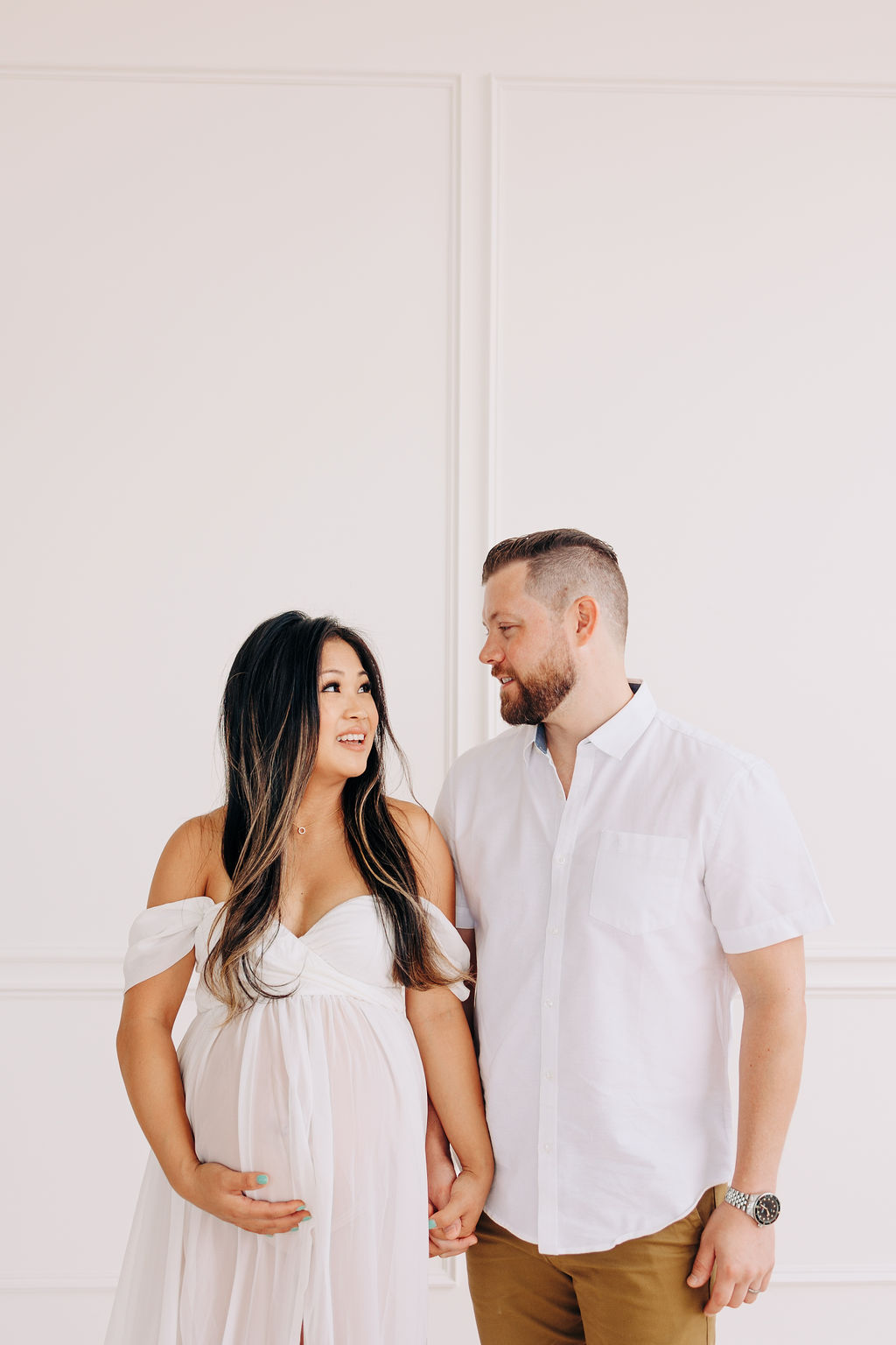 A mom to be holds her bump and her partner's hand while they stand in a studio against a white wall and looking at each other prenatal yoga in houston