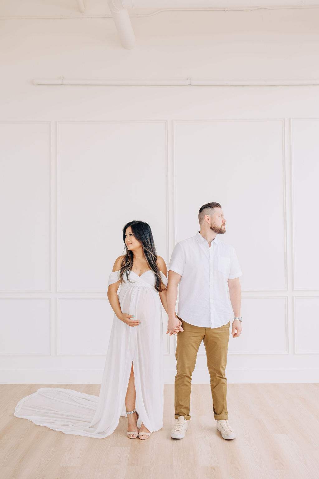 Expecting parents stand together in a studio holding hands and looking away from each other as mom wears a long white maternity gown and dad a white shirt and khakis prenatal yoga in houston
