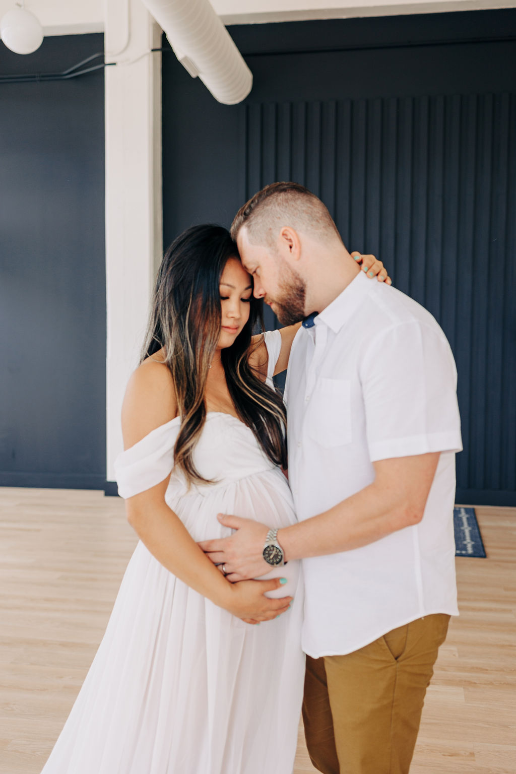 A mom to be stands with her husband with hands on the bump in a studio while wearing a white shirt and a long white maternity gown prenatal yoga in houston