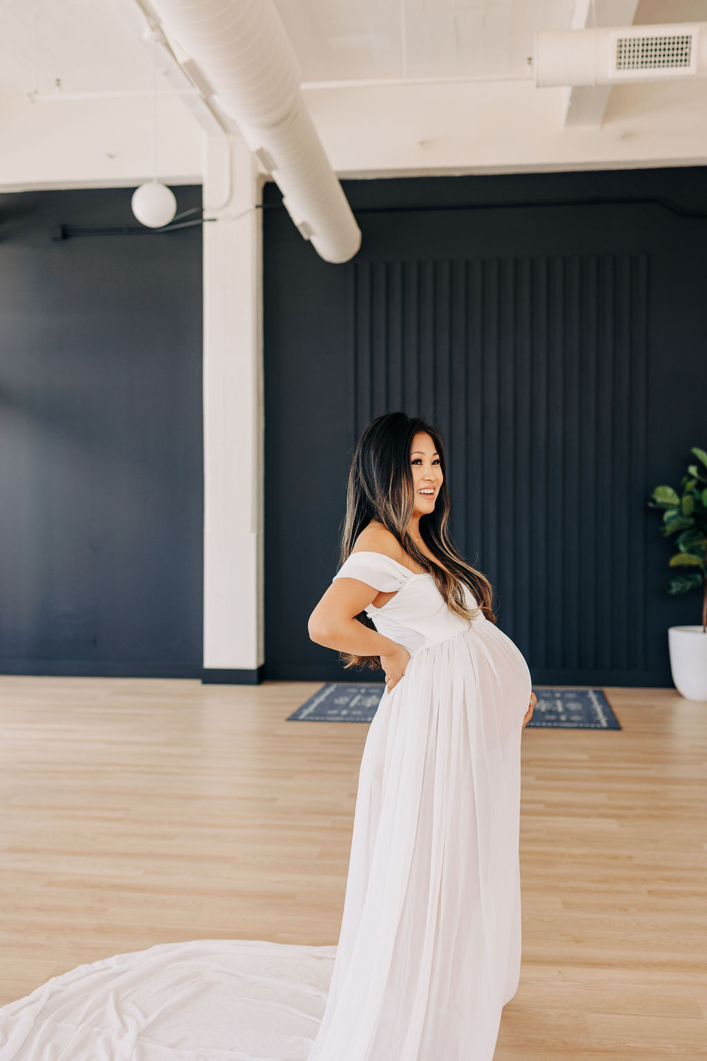 A mother to be in a long white maternity gown laughs while standing in a studio with wood floor 