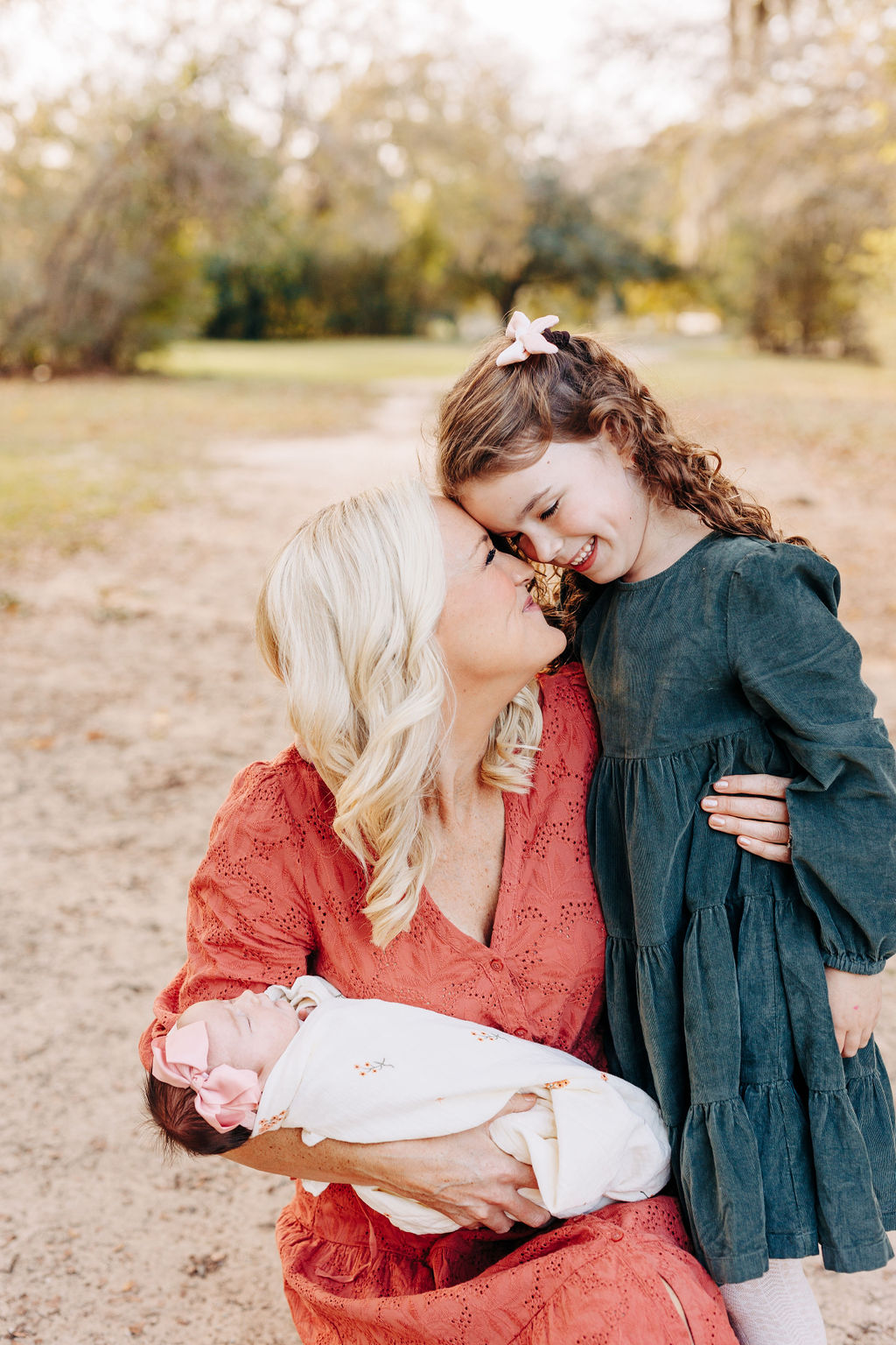 A happy mom hugs her young daughter while kneeling in a park path and holding her newborn baby girl in the other arm before family friendly restaurants in houston