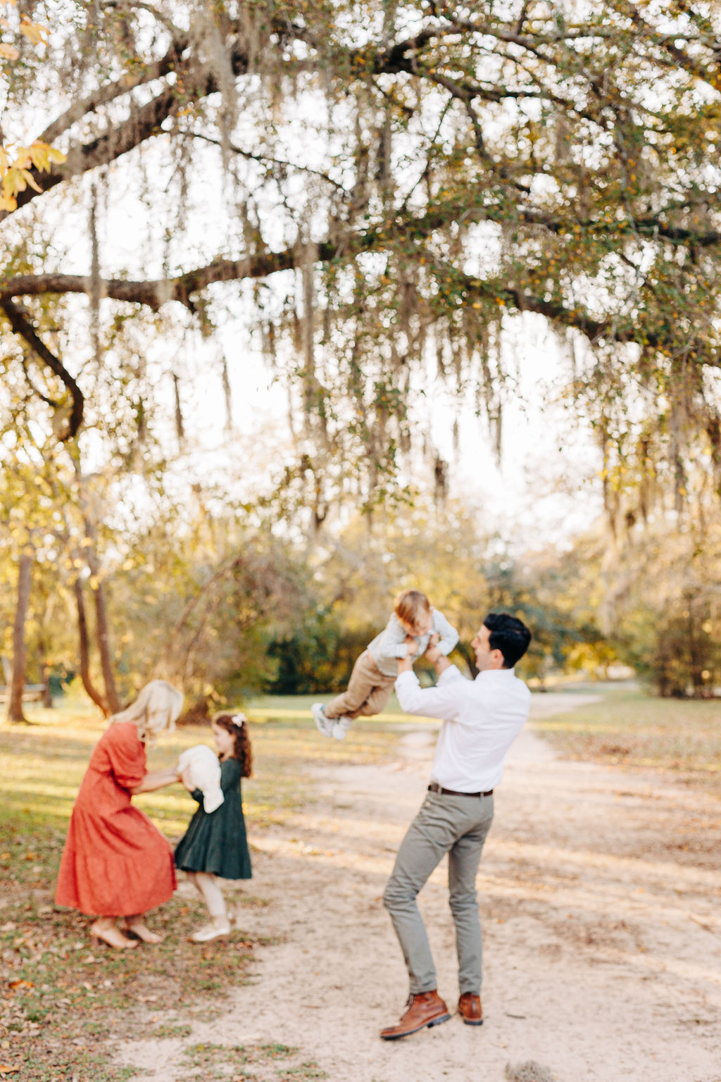A father lifts and spins with his toddler son while mom helps their daughter hold a newborn baby in a park