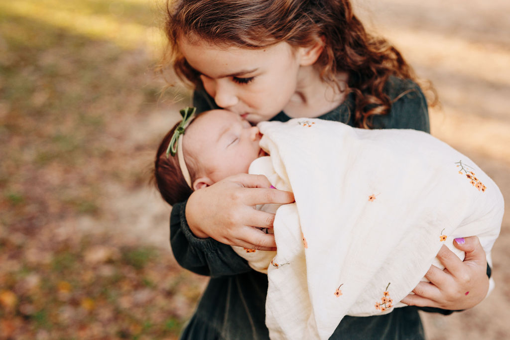 A young girl in a green dress kisses the forehead of her sleeping newborn baby sister in a park