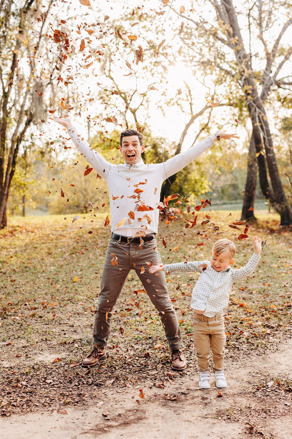 A happy dad throws leaves up in the air while playing with his toddler son in a park