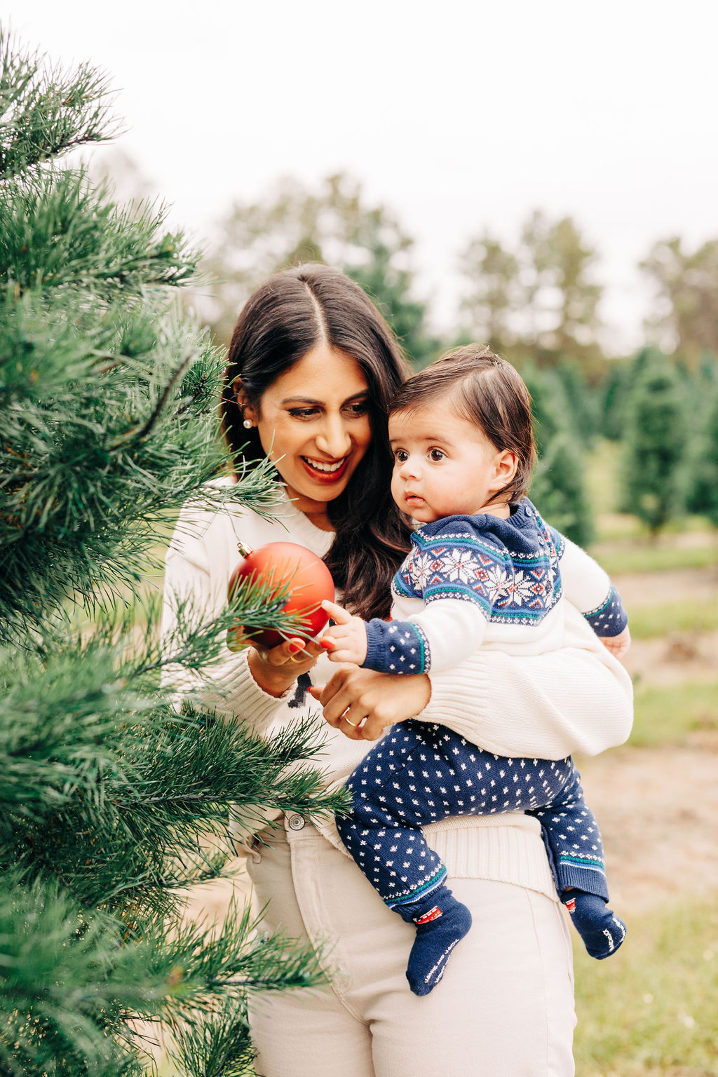 A mother shows her toddler baby a christmas ornament hanging on a tree in a farm