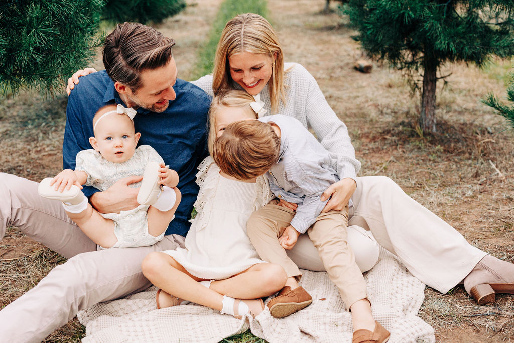 A mom and dad sit on a picnic blanket playing with their three young children in houston christmas tree farms