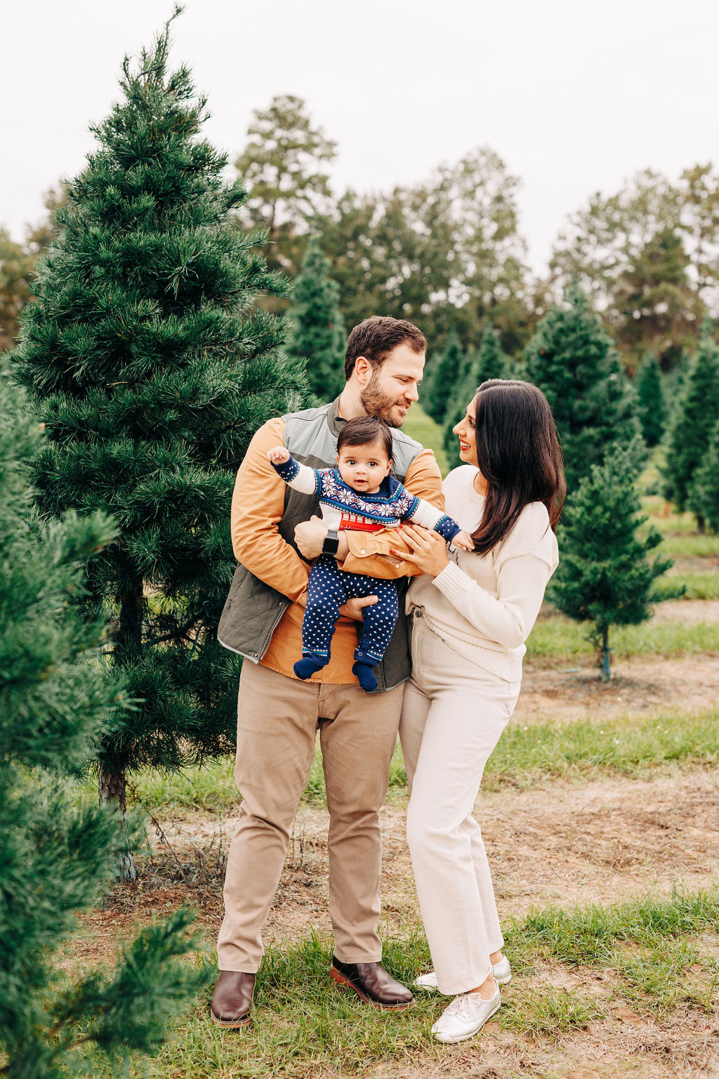 Excited parents smile at each other while holding their toddler in a christmas tree farm