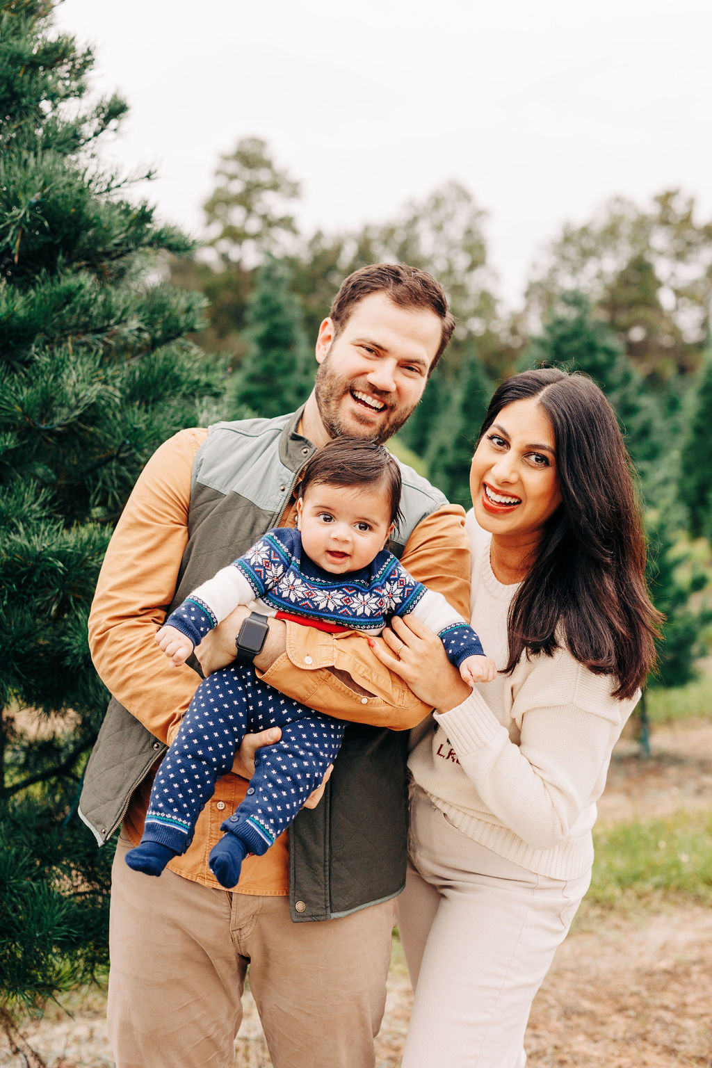 Happy parents laugh and play with their toddler in a christmas sweater onesie among christmas trees