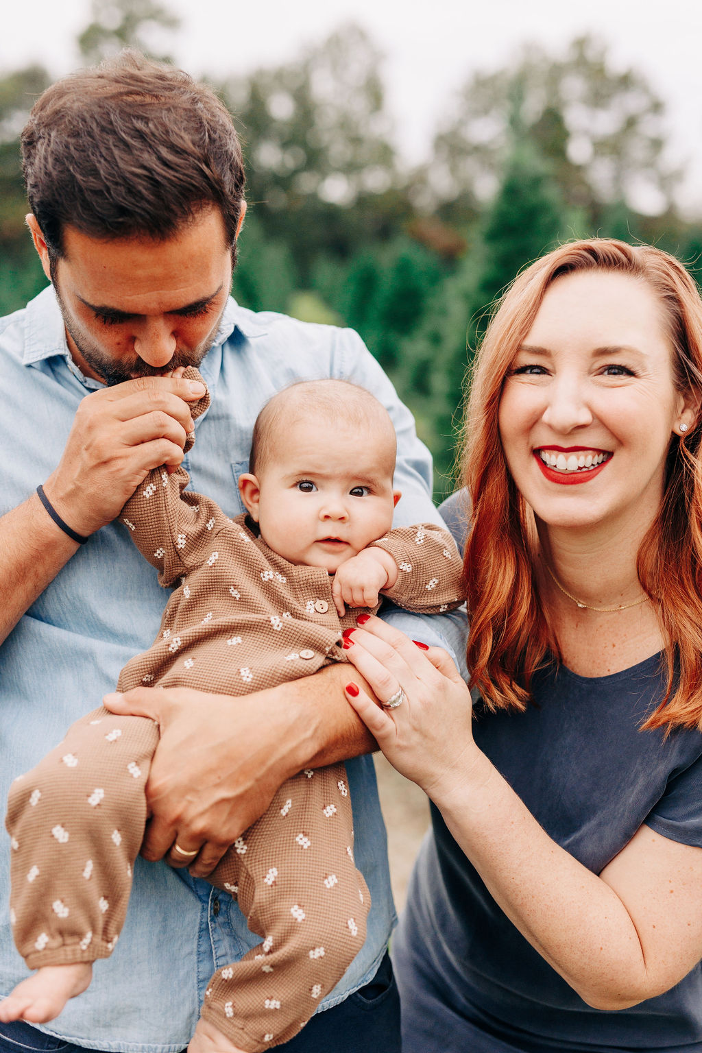 A father kisses the hand of his infant child in a brown onesie while standing with mom