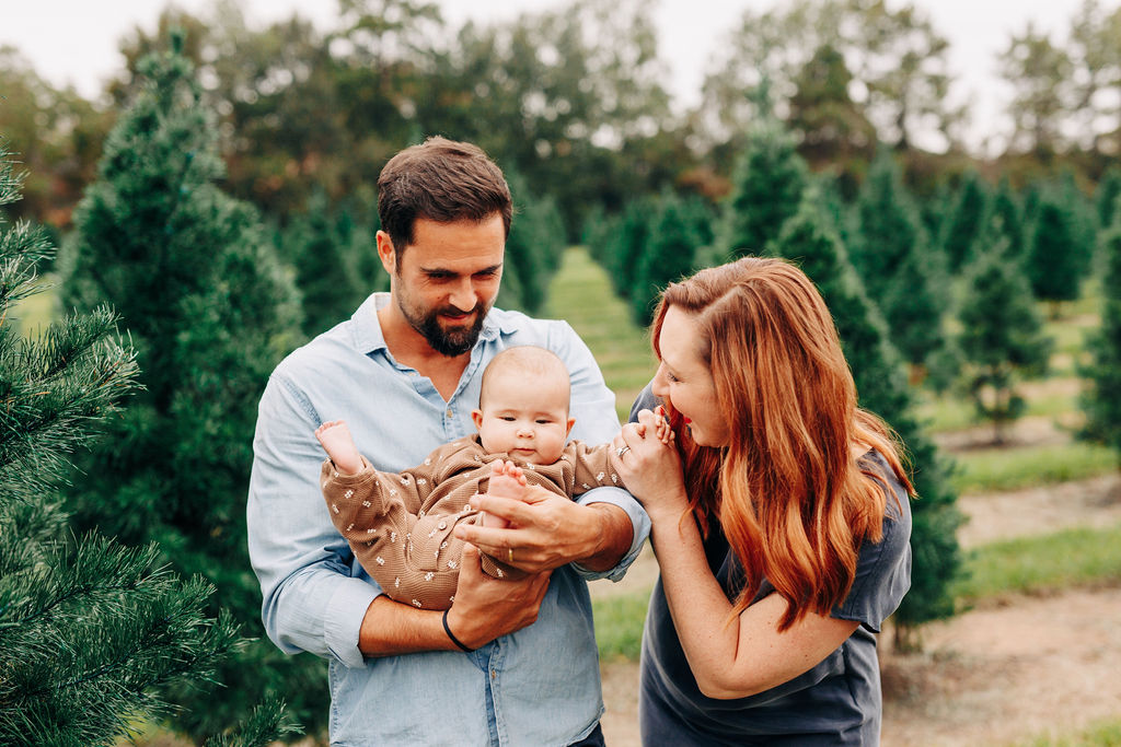 A mother plays with the hand of her infant baby boy being held by dad in houston christmas tree farms