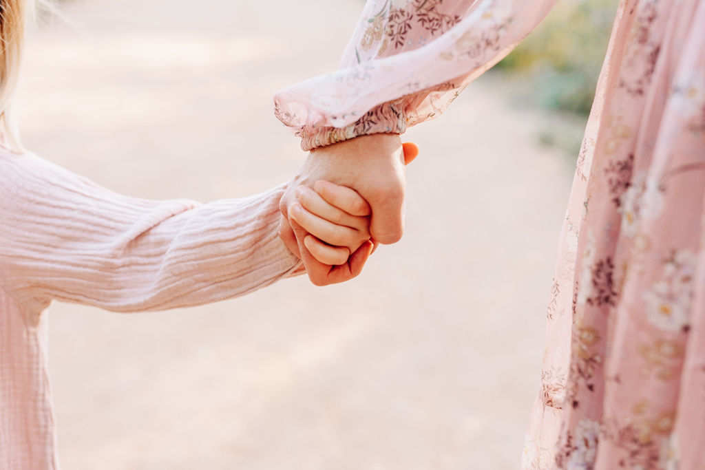 Details of a mom holding hands wit her toddler daughter in pink dresses in a park