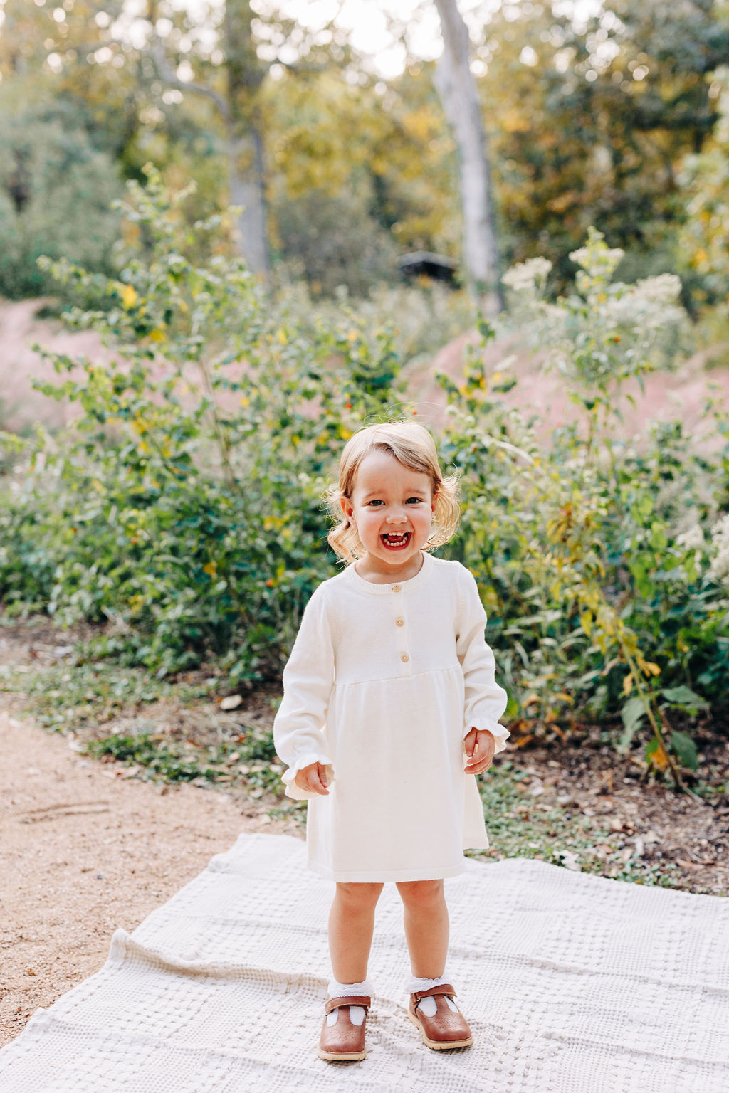 A toddler girl in a white dress laughs while standing on a picnic blanket in a park path